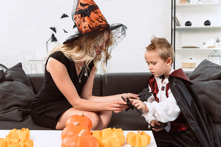 mother and little son in halloween costumes with black paper bats on sofa at homeの写真素材