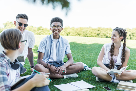 happy multiethnic teenage students sitting on grass and studying together in parkの写真素材