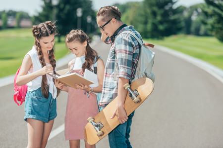 teenage students with backpacks and skateboard standing and reading book together in parkの写真素材