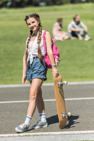 beautiful teenage girl with backpack standing with skateboard and smiling at camera in parkの写真素材