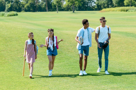 multiethnic teenage students with books, backpacks and sport equipment walking together in parkの写真素材