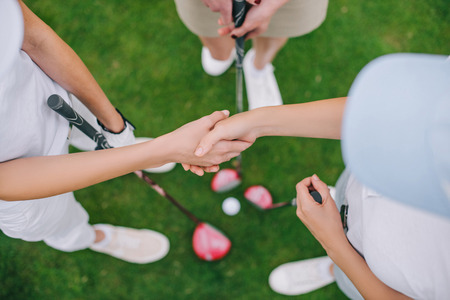 overhead view of female golf players with golf clubs shaking hands while standing on green lawnの写真素材
