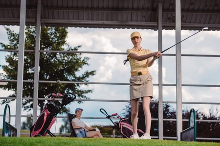 selective focus of female golf player playing golf while friend resting behind at golf courseの写真素材
