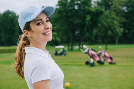side view of smiling woman in polo and cap looking at camera at golf courseの写真素材
