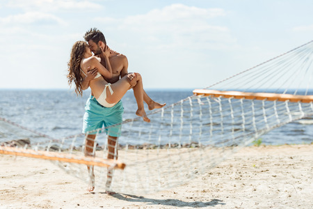 man holding girlfriend on hands and kissing her on beach, with hammock on foregroundの写真素材