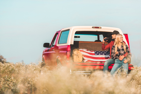 relaxed young couple sitting together in car trunk and drinking beer in flower fieldの写真素材