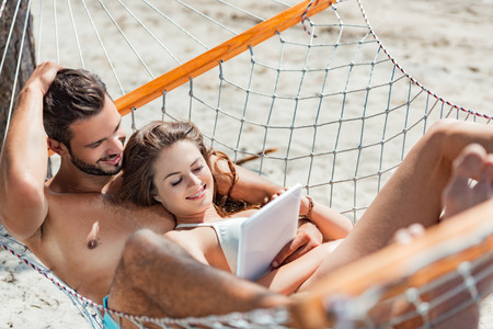 smiling couple using digital tablet while relaxing on hammock on beachの写真素材