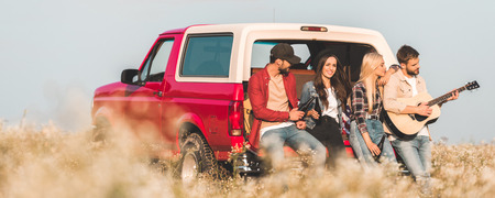 group of happy young friends drinking beer and playing guitar while relaxing in car trunk in flower...