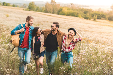 group of happy young friends embracing and walking by field together during trip
