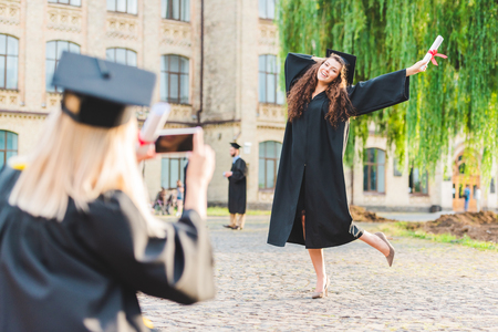 Partial view of woman taking picture of classmate with diploma during graduation near universityの写真素材