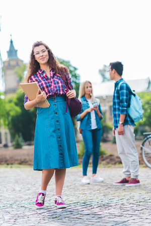 Selective focus of smiling student with notebook and multicultural classmates behind on streetの写真素材