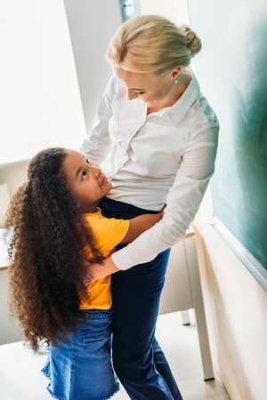 Adorable African american schoolgirl embracing her teacher at classroomの写真素材