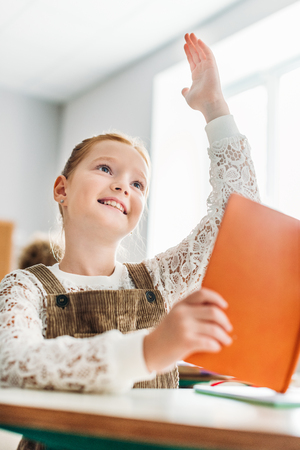 Smiling little schoolgirl with book raising hand during lessonの写真素材