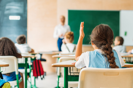 Back view of schoolgirl raising hand to answer teachers question during lesson