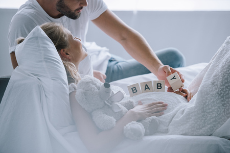 partial view of man putting wooden blocks with baby lettering on belly of pregnant wife with teddy...