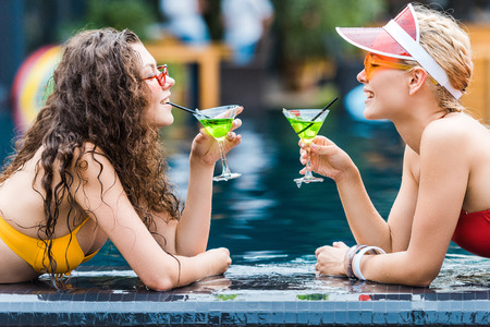 side view of two attractive smiling female friends drinking cocktails on poolsideの写真素材
