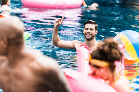 smiling man with raised finger in swimming pool talking to friendsの写真素材
