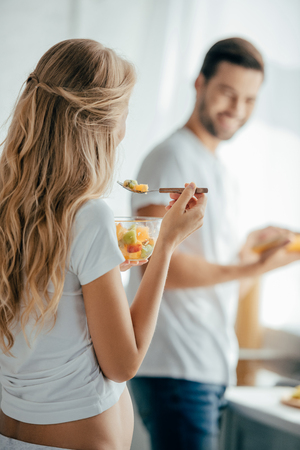 selective focus of pregnant woman eating fruits salad while smiling husband standing at counter in kitchenの写真素材