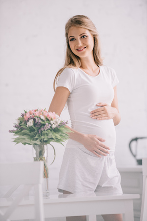 happy pregnant woman standing at table with bouquet of flowers in vase at homeの写真素材