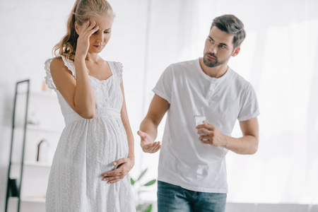 pregnant woman in white nightie having headache while husband giving medicines and glass of water to her at homeの写真素材