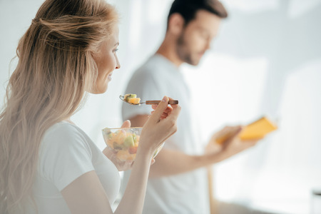selective focus of smiling pregnant woman eating fruits salad while husband standing at counter in kitchenの写真素材