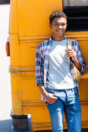 happy teen african american schoolboy leaning back on school bus