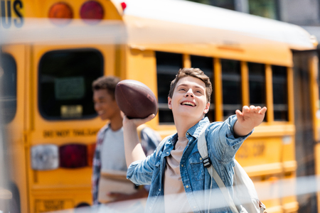 happy teen schoolboy throwing american football ball in front of school bus with blurred classmate walking on backgroundの写真素材