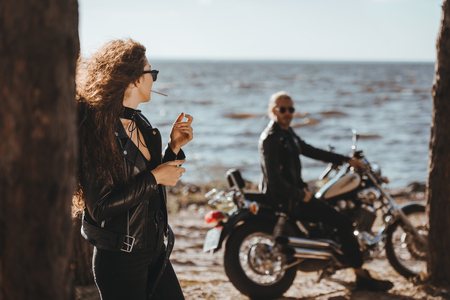 selective focus of woman smoking cigarette and looking at her boyfriend on motorbike on seashoreの写真素材