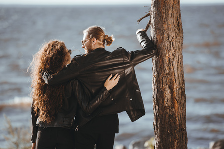 back view of couple in black leather jackets hugging and looking at each other on seashoreの写真素材