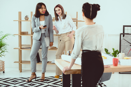 Back view of young businesswoman standing at table and smiling colleagues talking behindの写真素材