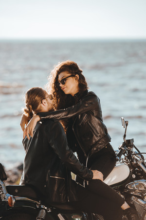 couple of bikers in black leather jackets embracing on classic motorcycle on seashoreの写真素材