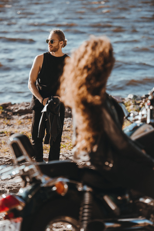 selective focus of girl sitting on motorcycle and looking at boyfriend walking on seashoreの写真素材
