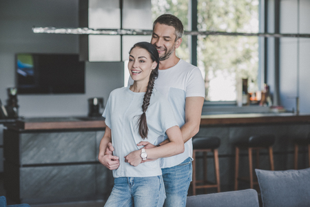 happy man hugging girlfriend from behind in kitchen at homeの写真素材