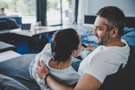 smiling man with remote controller embracing girlfriend and watching tv on sofa at homeの写真素材