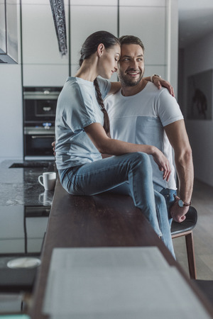 side view of attractive woman sitting on kitchen counter and embracing boyfriend at homeの写真素材