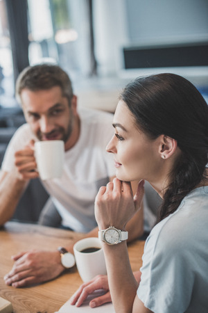 side view of young woman sitting at table with boyfriend while he drinking coffee at homeの写真素材