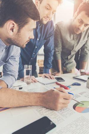 Three young businessmen leaning at table and working at project together, business teamwork conceptの写真素材