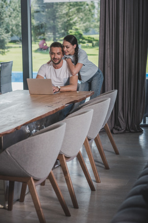 happy young woman embracing boyfriend while he using laptop at homeの写真素材