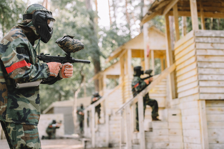 side view of paintballer in camouflage and protective mask aiming by marker gun while his team standing behind outdoorsの写真素材