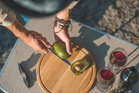 cropped view of man cutting avocado on wooden board on table with wine