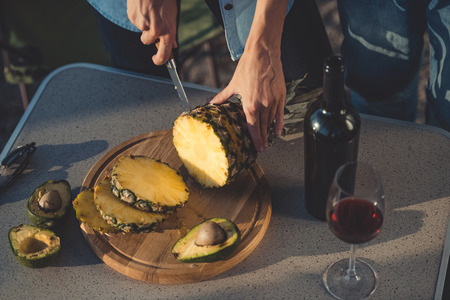 cropped view of woman cutting pineapple and avocados on wooden board near bottle of red wine