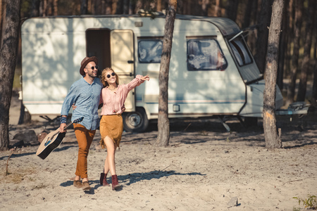 young couple in sunglasses hugging and walking with acoustic guitar near trailer in forest, girl showing somethingの写真素材