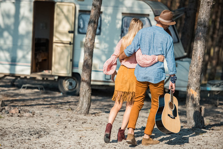 rear view of hippie couple hugging and walking with guitar near trailerの写真素材