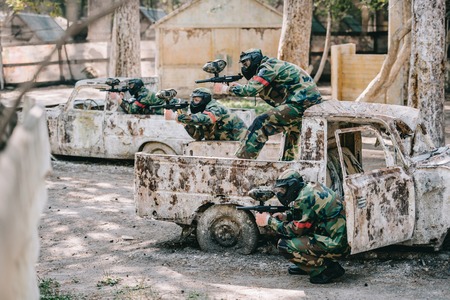paintball team in camouflage and goggle masks aiming with marker guns from broken cars outdoorsの写真素材