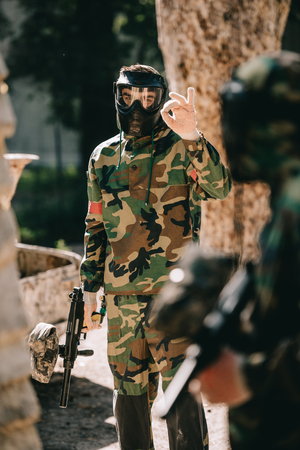 male paintball player in camouflage and goggle mask showing ok sign to his team outdoorsの写真素材