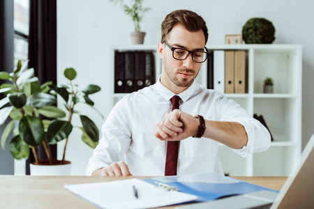 handsome businessman looking at wristwatch while sitting at table with documents