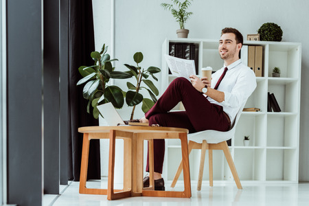stylish businessman with coffee reading newspaper in modern office with laptopの写真素材