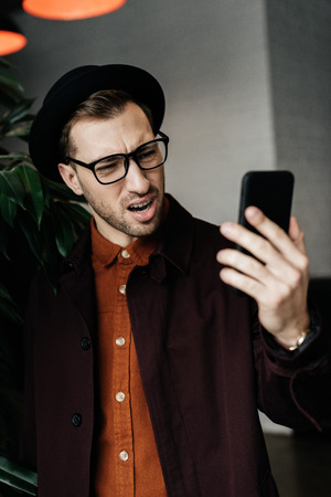 handsome stressed man in hat and glasses looking at smartphoneの写真素材