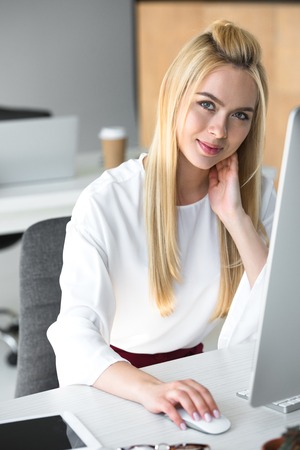 beautiful young woman using desktop computer and smiling at camera in officeの写真素材