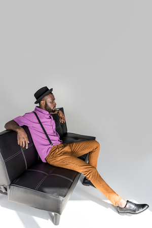 side view of african american stylish man in hat resting on black sofa on grey backdropの写真素材